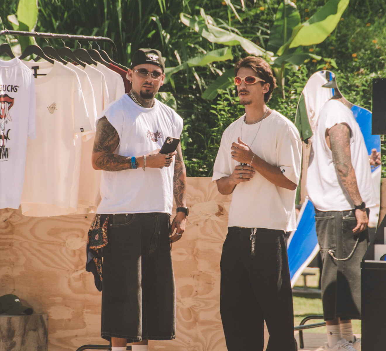 Dois jovens no estande da Fivebucks no SEAS 10 anos — plataforma de madeira com araras e looks da marca