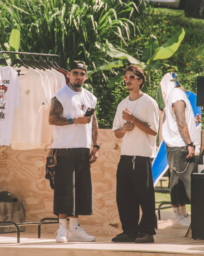 Dois jovens no estande da Fivebucks no SEAS 10 anos — plataforma de madeira com araras e looks da marca